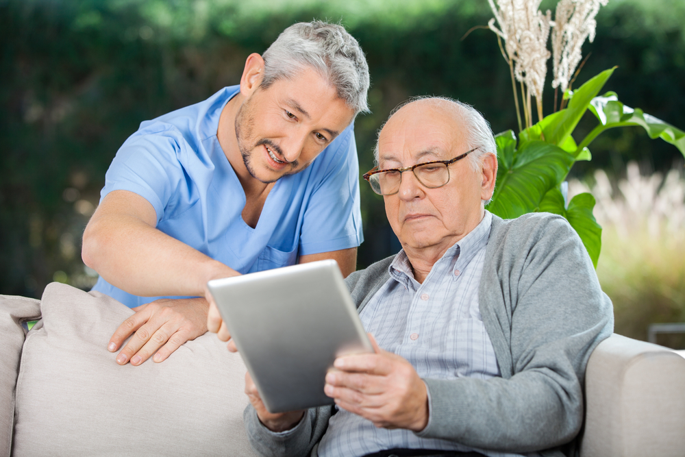 An elderly man holds an iPad while an in-home nurse assists him with what he is viewing on his screen.