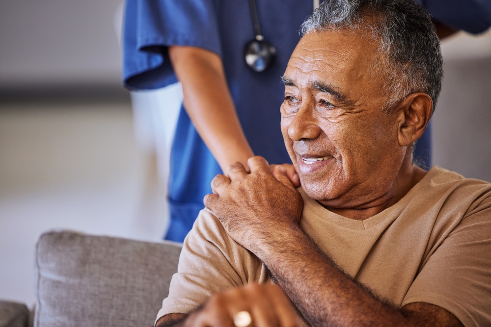 Senior man sitting down with a caregiver behind him resting their hand on his shoulder.