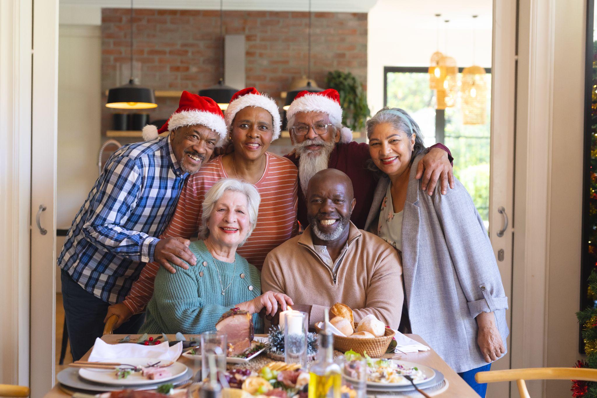 Diverse seniors dressed in Santa hats gathered around a table smiling at the camera.