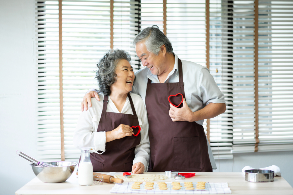 Senior couple with aprons on standing in the kitchen holding cookie-cutters in the shape of hearts over the chests.